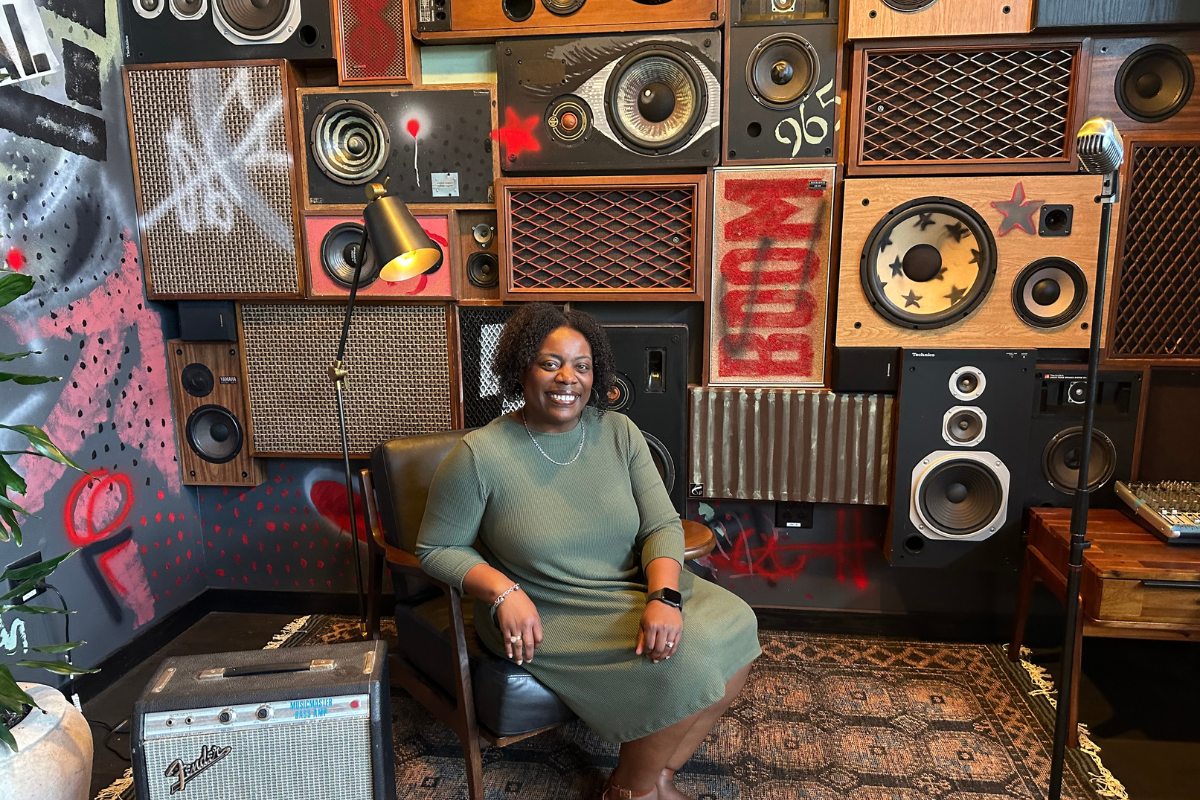 Cherith smiling in a leather chair in front of a graffiti-covered wall of vintage speakers and a Fender amp.