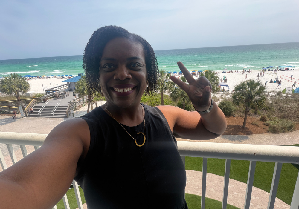 Cherith Fluker takes a selfie on a hotel balcony, flashing a peace sign with turquoise Gulf water, white sand beach, and palm trees visible behind her.
