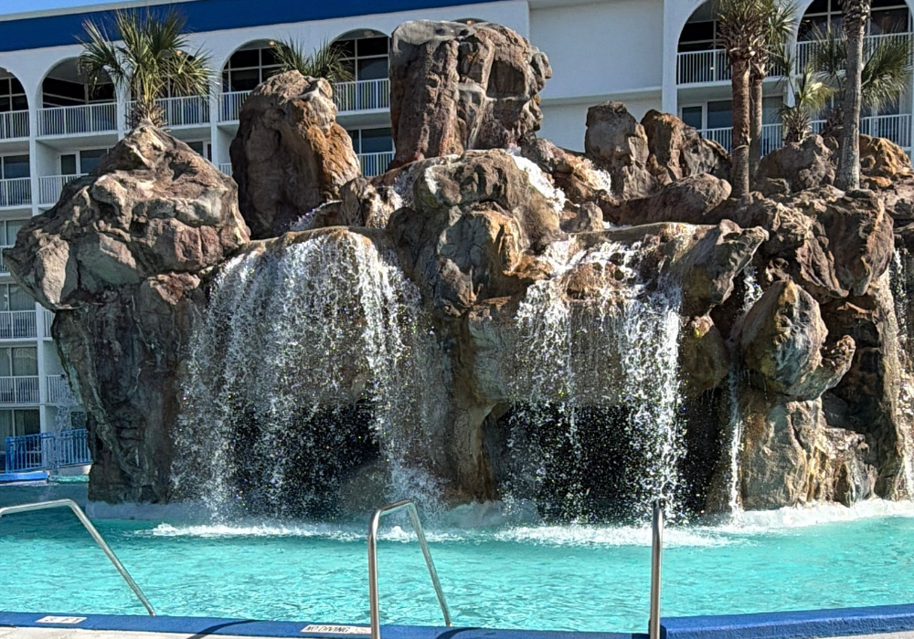 A large grotto-style pool feature at The Island Resort, with water cascading down multiple levels of rugged artificial rock formations into a bright turquoise pool, flanked by palm trees and hotel balconies in the background.
