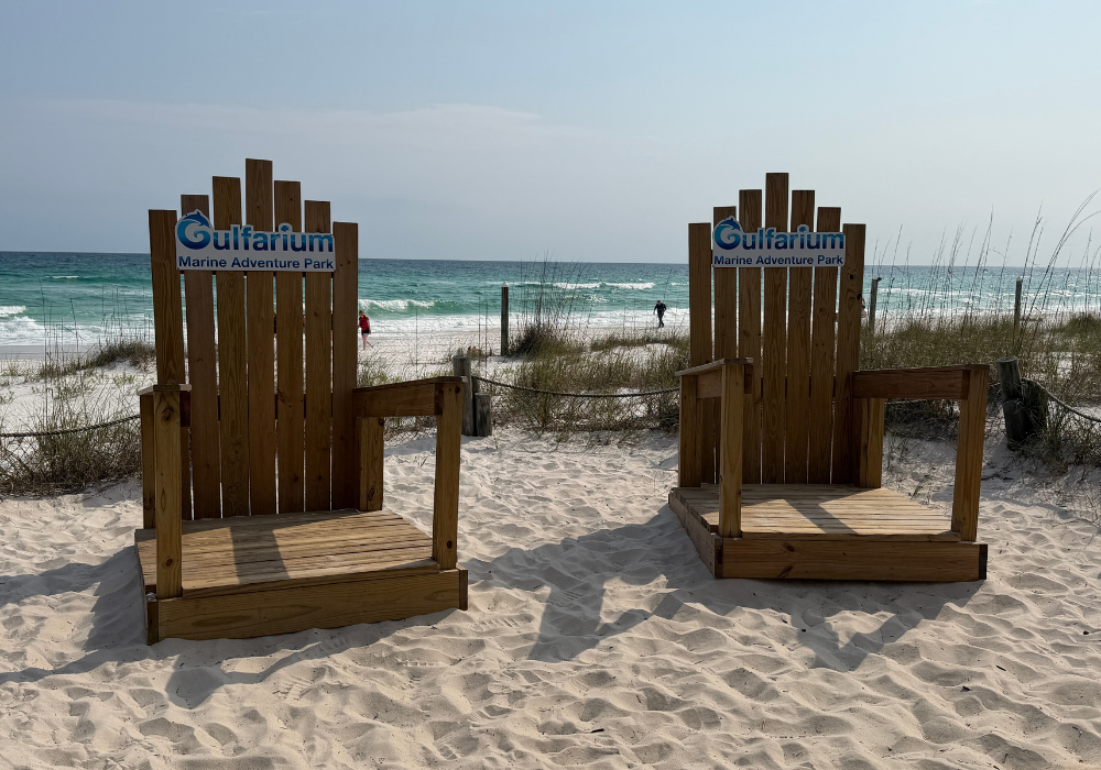 Two oversized wooden Adirondack chairs branded with the Gulfarium Marine Adventure Park logo, positioned on a white sand beach with sea oats and the emerald Gulf of Mexico visible in the background.

