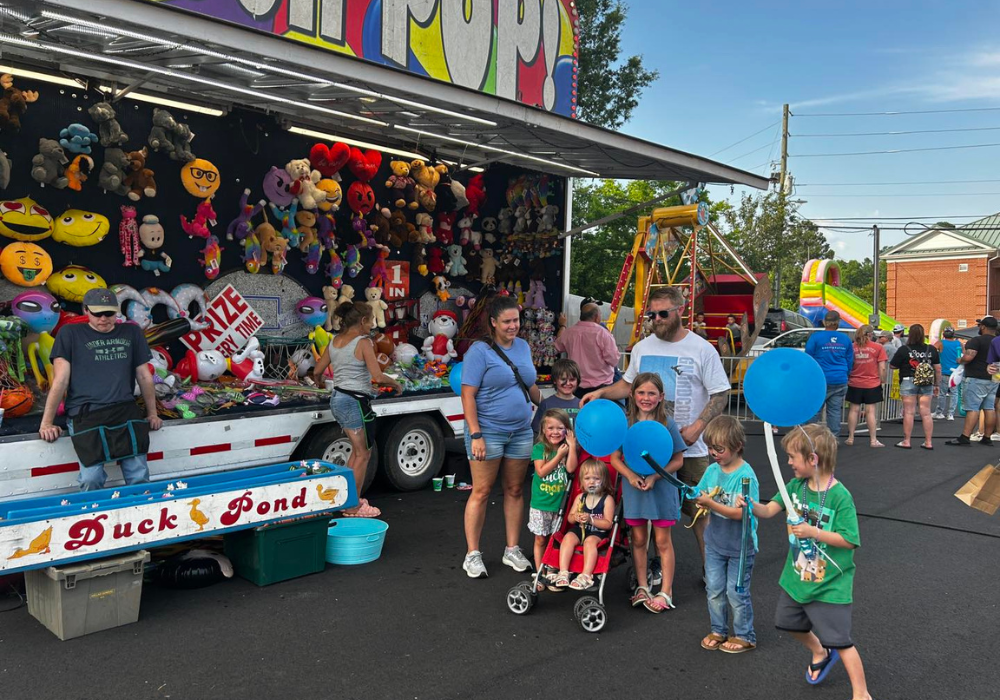 A family with young children stands near a carnival-style Duck Pond game and prize booth in the kids' zone at the Pell City Block Party, with inflatables and rides visible in the background.