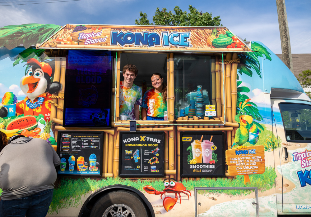 Two smiling Kona Ice employees lean out of a colorfully decorated tropical shaved ice truck at the Pell City Block Party in Oxford, Alabama.