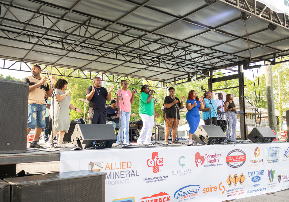 A large gospel and R&B ensemble performs on an outdoor stage at the Pell City Block Party, with sponsor banners visible along the front of the stage.