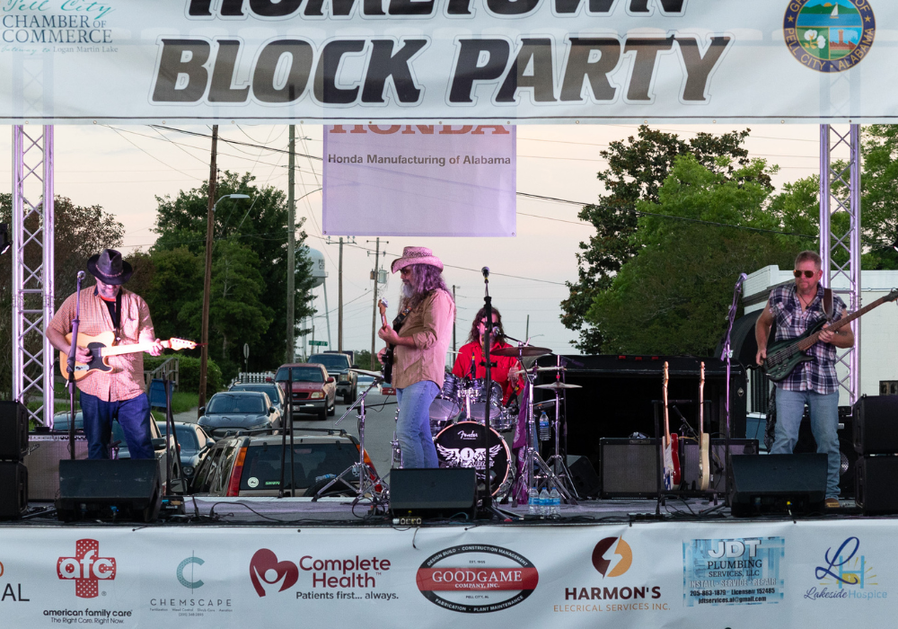 Four-member band performing on an outdoor stage at a town block party, with a large 'Hometown Block Party' banner above and sponsor banners along the front.