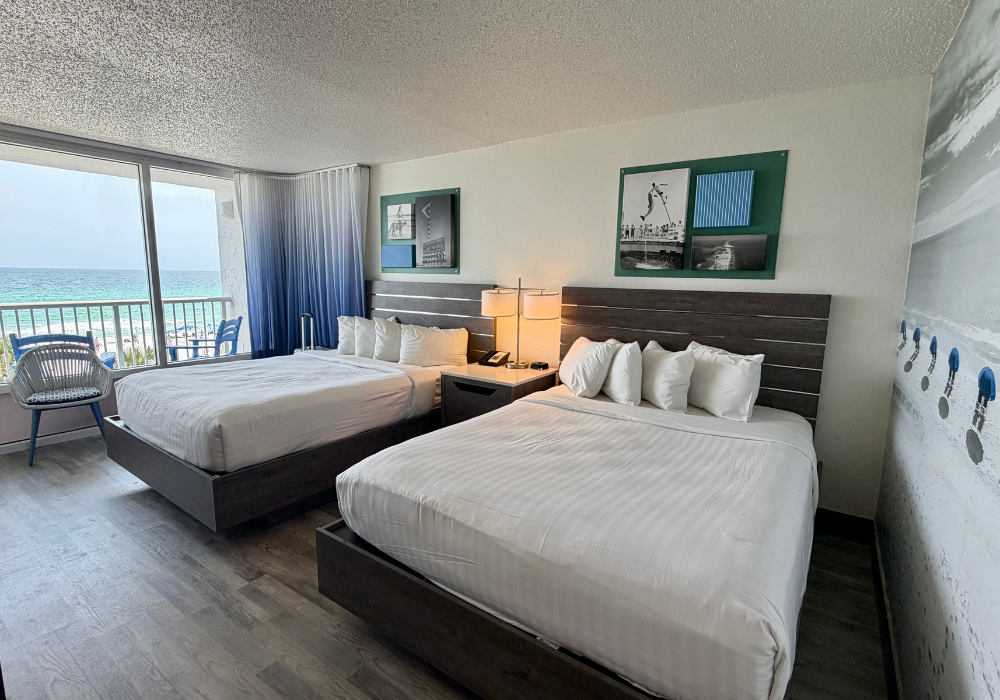 A renovated hotel room with two queen beds dressed in white linens, wood-plank flooring, dark wood headboards, coastal-themed black and white artwork in teal frames, and a sliding glass door opening to a balcony with a direct view of the Gulf of Mexico.
