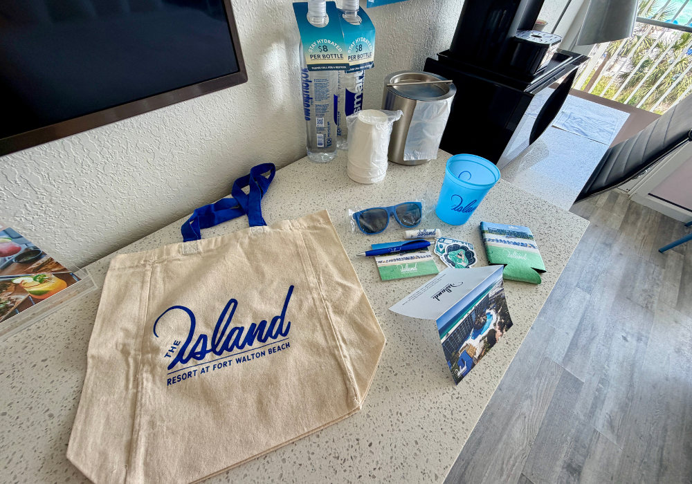A welcome amenity spread on a hotel room counter including a canvas tote bag branded "The Island Resort at Fort Walton Beach," sunglasses, a tumbler cup, koozies, stickers, pens, brochures, and two water bottles.
