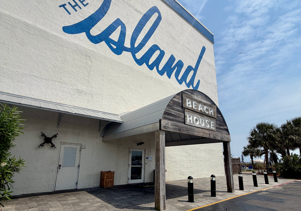 Side entrance of The Island Resort featuring a weathered wood canopy sign reading "Beach House" beneath large blue script lettering spelling "The Island" painted on the white exterior wall of the building.
