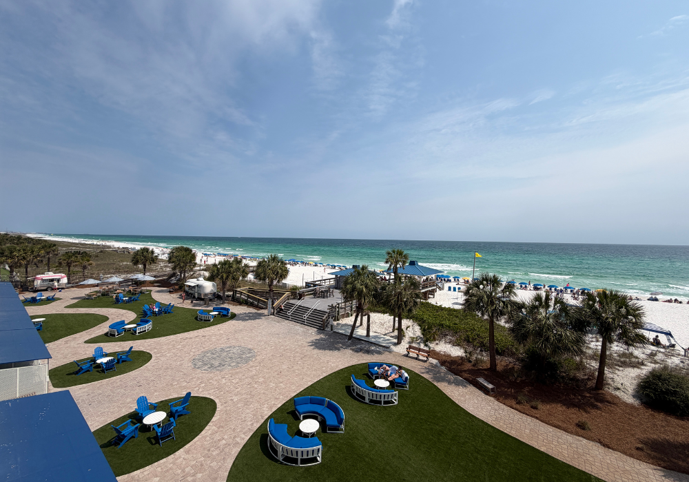 Elevated view from a hotel balcony showing a landscaped courtyard with blue Adirondack chairs, circular seating areas on green grass, palm trees, a beachside deck, and the emerald green Gulf of Mexico stretching to the horizon.