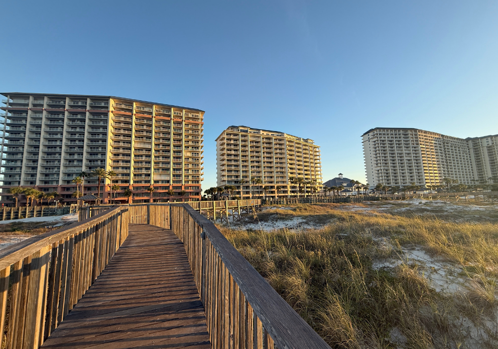 A wooden boardwalk leads through the dunes to the beach at The Beach Club Resort in Gulf Shores, Alabama, with the towers glowing in late afternoon sun.